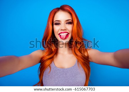 Portrait of stylish funny happy carefree girl with ginger hair,cute face and big breast make selfie photo and put out tongue while standing on blue background