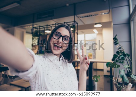 Portrait of excited cheerful smiling young pretty woman in spectacles making selfie photo and showing v-sign with two fingers Royalty-Free Stock Photo #615099977