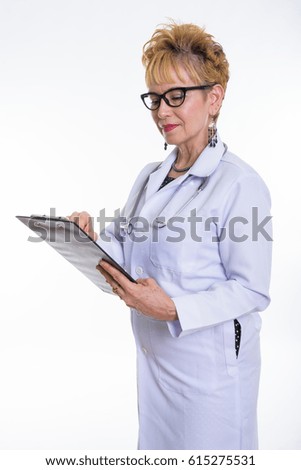 Studio shot of senior Asian woman doctor standing while reading on clipboard and wearing eyeglasses against white background