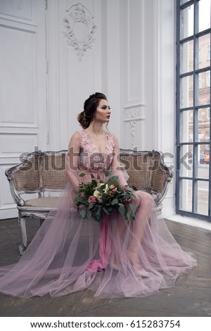 bride's morning. Young girl posing in pink sheer nightie in a large white bright room in a beautiful old chair with a bouquet of flowers