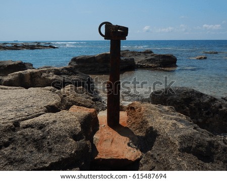 Rusted fishing boat winch standing amongst rocks on Vardas beach with sea and rocky outcrop in background.