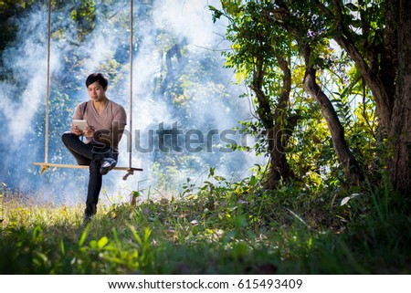 Young man is playing tablet and an old camera while sitting on a swing in the forest.