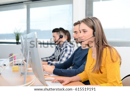 portrait of a beautiful and cheerful young woman telephone operator with headset working on desktop computer in row customer service call support helpline business center with teamworker in background