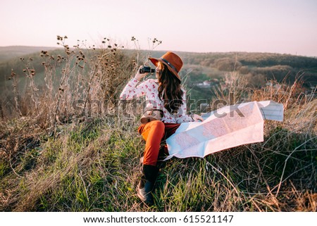 Traveler beautiful girl with a hat looking at the side. map, holding in her hand binoculars at sunset sitting on a background of mountains. Concept photo travel, adventure