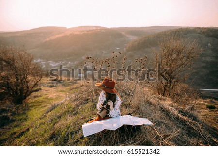 Traveler beautiful girl with a hat looking at a map holding in her hand binoculars at sunset sitting on a background of mountains. Concept photo travel, adventure
