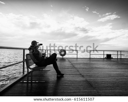 Man hiker on mole at sea. Tourist on wharf looking over sea to horizon. Sunny clear blue sky, smooth water level. Black and white photo