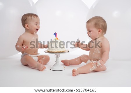 One year old twin boys wearing diapers and eating birthday cake. The cleaner of the two appears to be laughing at his messy brother.