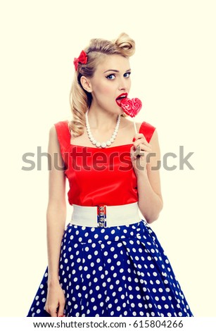 Portrait of woman eating heart shape lollipop dressed in pinup style dress in polka dot. Caucasian blond model posing in retro fashion and vintage concept studio shoot.