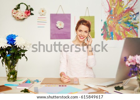 Beautiful fashion designer with charming smile distracted from making sketch due to telephone call, abstract painting and flower decorations hanging on wall