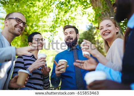 Cheerful group of colleagues having coffee break: they standing with paper cups in sunny spring park and chatting animatedly, low angle view