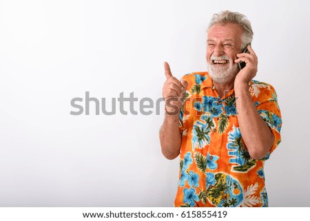 Studio shot of thoughtful happy senior bearded tourist man smiling and laughing while talking on mobile phone and pointing finger against white background