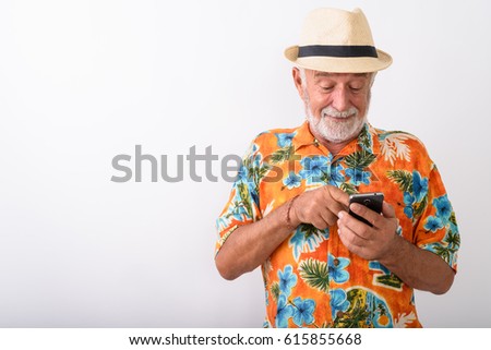Studio shot of happy senior bearded tourist man smiling while using mobile phone and wearing hat against white background