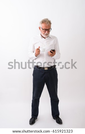 Full body shot of handsome senior bearded man standing while using mobile phone and holding coffee cup against white background