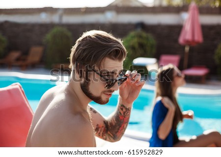 Young handsome man in sunglasses sunbathing, sitting near swimming pool.