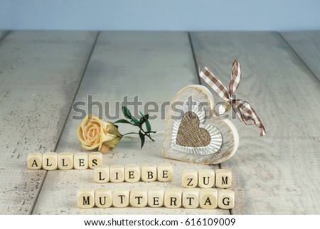 The German words for All Love for Mother's Day in wooden letters, with a rustic wooden heart and a dry flower in the background