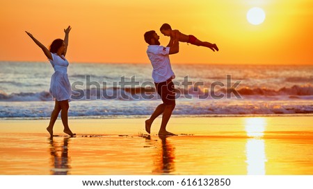 Happy family - father, mother, baby son walk with fun along edge of sunset sea surf on black sand beach. Active parents and people outdoor activity on summer vacations with children on Bali island