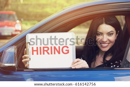 Happy woman sitting inside new car showing white card with hiring sign message on a dealership lot background 