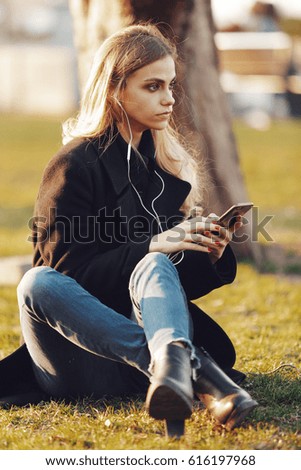 Young beautiful girl is holding a smartphone and listening to music on headphones while sitting on the grass in a megapolis park