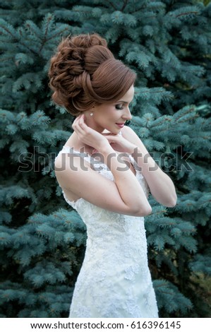 Young model with bridal hairstyle and make-up, in bridal white dress. A spruce tree is on the background.