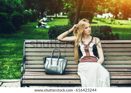 Attractive pensive blonde woman with long hair sitting on park bench with bag near her, adult beautiful lady thoughtfully looking aside while waiting her boyfriend in city parkland on sunny summer day