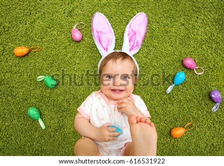 Portrait of happy smiling baby in bunny ears with Easter eggs lying down on a green rug, top view. Kids celebrate Easter. 