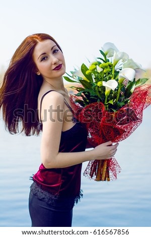 Beautiful red-haired elegant young lady with pale skin with a bouquet of white callas near the blue water with evening make-up lipstick of marsala color and arrows on the eyes at sunny day