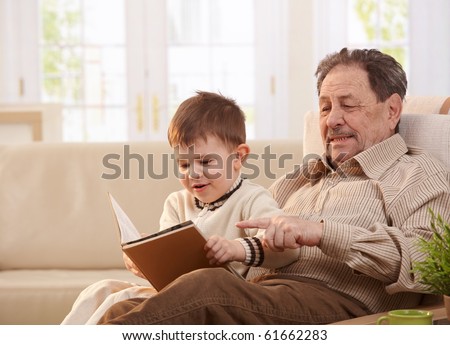 Grandfather sitting in armchair at home, holding his grandson who looking pictures in book.?
