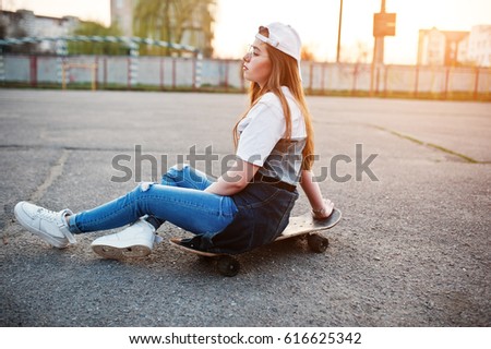 Young teenage urban girl with skateboard, wear on glasses, cap and ripped jeans at the yard sports ground on sunset.