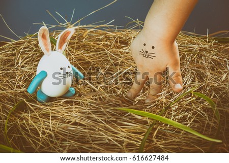 Funny easter rabbit and child hand with cute face. Nest from dry grass with green leaves. Face painted on a child's hand. Toned.