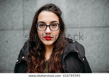 Fashion concept: beautiful young girl with long hair, glasses, red lips standing near modern wall wearing in green suit and grey jeans. She wanted talking on phone. Color