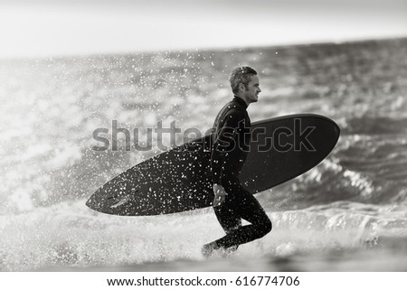 Surfer silhouette of a man holding his surf board, he is running on the beach, he is wearing wetsuit. Black and white picture
