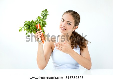 Beautiful young woman holding vegetables and fresh juice, sitting at a white table in light clothes on the light background