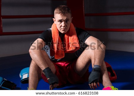 Young male boxer with a towel around his neck sitting near lying boxing gloves and helmet in regular boxing ring in a gym