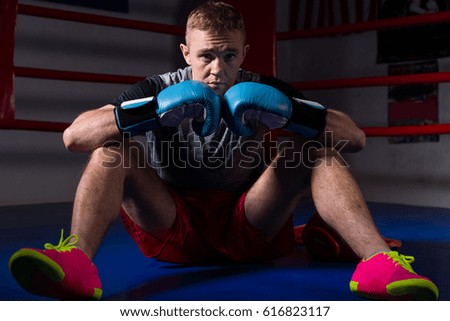 Young male boxer in boxing gloves sitting in regular boxing ring in a gym