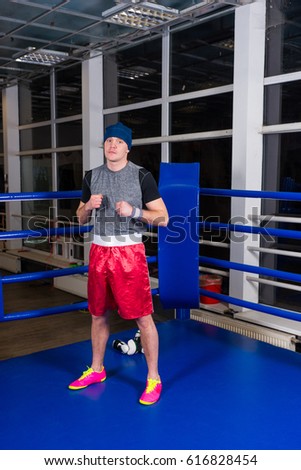 Athletic male boxer standing in a regular boxing ring surrounded by ropes in a gym