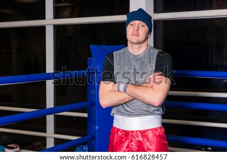 Athletic boxer with arms across standing in a regular boxing ring surrounded by ropes in a gym