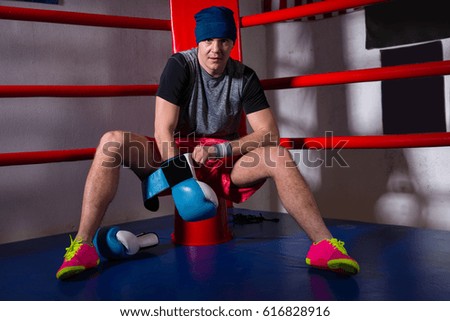 Spotive young male boxer sitting near red corner of a regular boxing ring and puts on boxing gloves in a gym