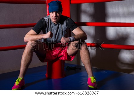 Athletic male boxer sitting near red corner of a regular boxing ring surrounded by ropes in a gym
