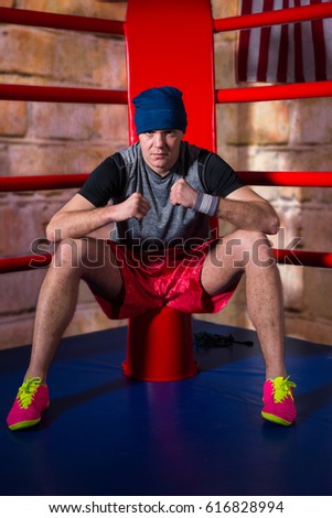 Athletic young male boxer sitting near red corner of a regular boxing ring surrounded by ropes in a gym
