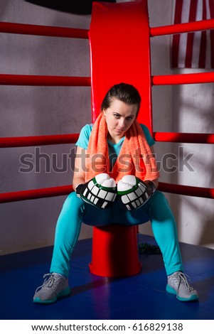 Young sporty female boxer with a towel around her neck sitting in regular boxing ring in a gym