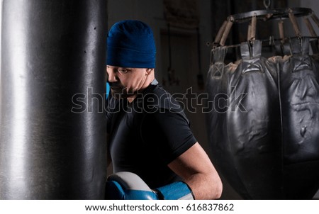 Male boxer with stern look in a hat and boxing gloves training with boxing punching bag in a gym