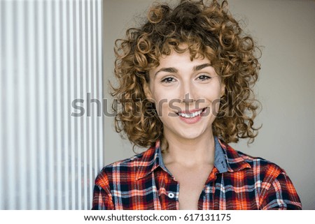 young natural curly woman smiling to camera casual dressed in a checkered shirt inside a house