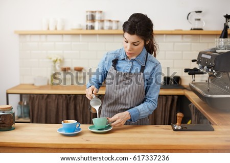 Beautiful female barista pours milk, making two cappuccino, looking concentrated.