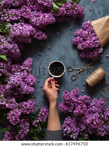 Hand holds a cup of morning coffee with spring lilac flowers branches blossoming on grey concrete background view from above. Flat lay underground style. Expensive colors. Creative design of flowers.