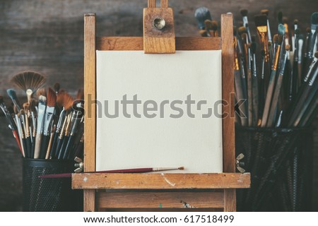 Artistic equipment in a artist studio: empty artist canvas on wooden easel and paint brushes Retro toned photo. Royalty-Free Stock Photo #617518499