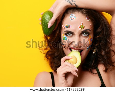 Closeup portrait of young happy beautiful fashion curly woman with cartoon fish stickers on face eating avocado on yellow background