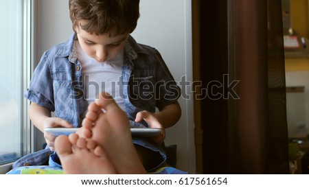 PORTRAIT: Cute little boy sits on a windowsill at home and touches a tablet PC