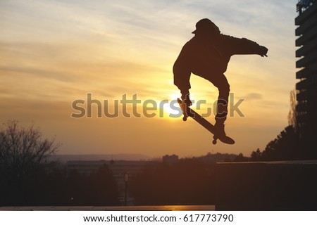 The teenager in a sweatshirt and a cap jumps with a board in the city against the backdrop of the urban sunset. Silhouette photo of extreme