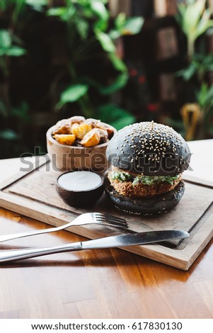 Vegetarian burger made with black charcoal bun with sesame served with potato wedges and sauce on wooden rustic table