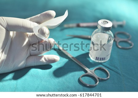 Hand holding silicone nasal implant in operating room with surgical equipment and medicine vial background.